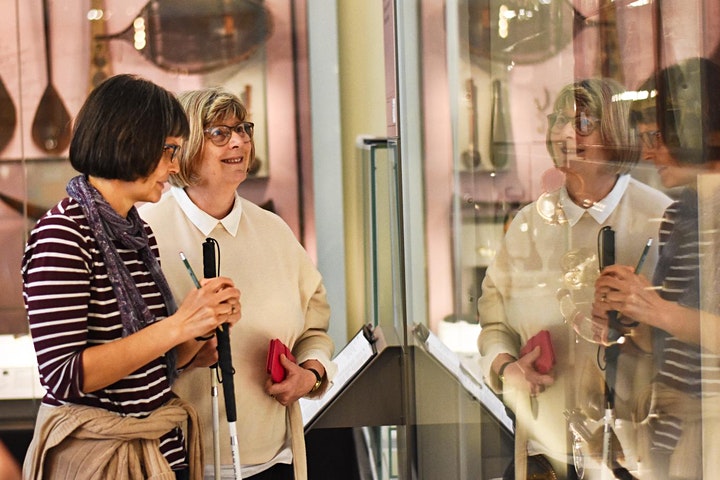 Zwei Frauen, eine mit und eine ohne Blindenstock scheun in einer Museum Vitrine ein