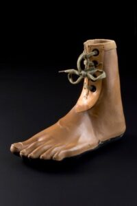 alt="A prosthetic foot made from brown wood and rubber, with thick laces at the ankle. Designed to appear barefooted."